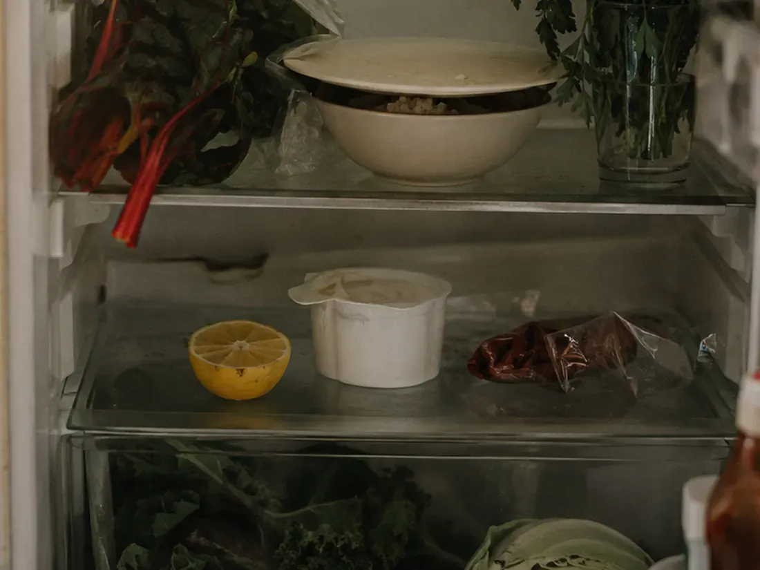 Étagères d'un réfrigérateur avec un reste dans un bol, un demi-citron, un pot de crème entamé et des bettes